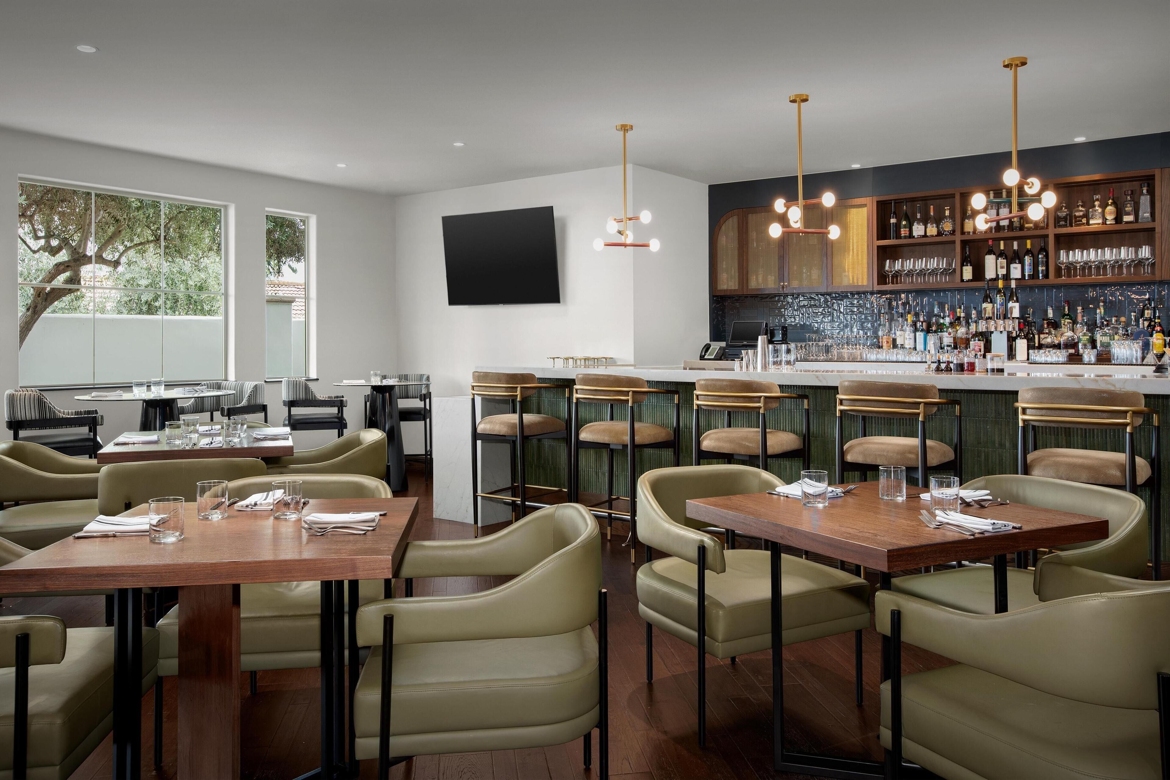 Een restaurant met hoge tafels en stoelen, een barruimte met flessen op de planken, en een groot raam met uitzicht op een boom.