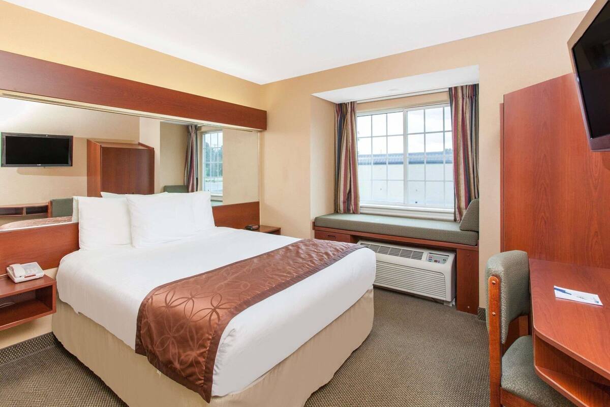 Habitación de hotel con cama, escritorio, silla, televisión, ventana con cortinas y aire acondicionado.