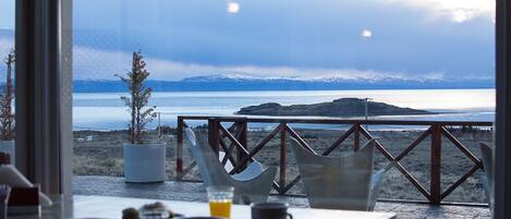 Zona de comedor con vistas a un lago y montañas. Hay sillas y mesas con tazas y vasos.
