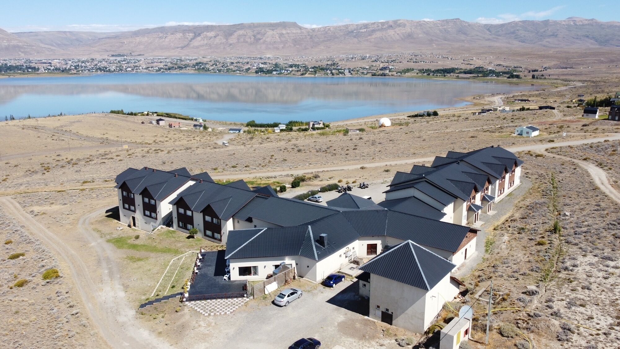 A large, modern residential complex with multiple houses, a central courtyard, and a view of a lake and mountains in the background.