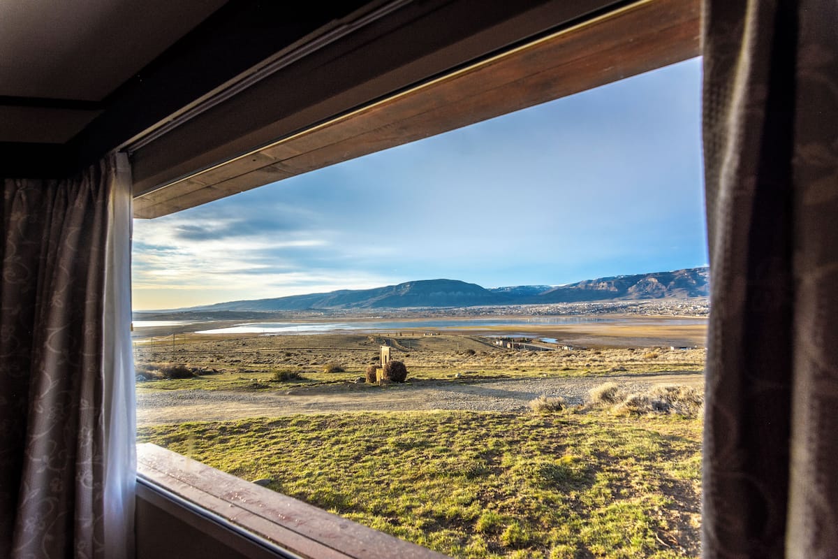 Vista da janela com cortinas, mostrando uma paisagem com um corpo d'água, colinas e uma construção ao longe.