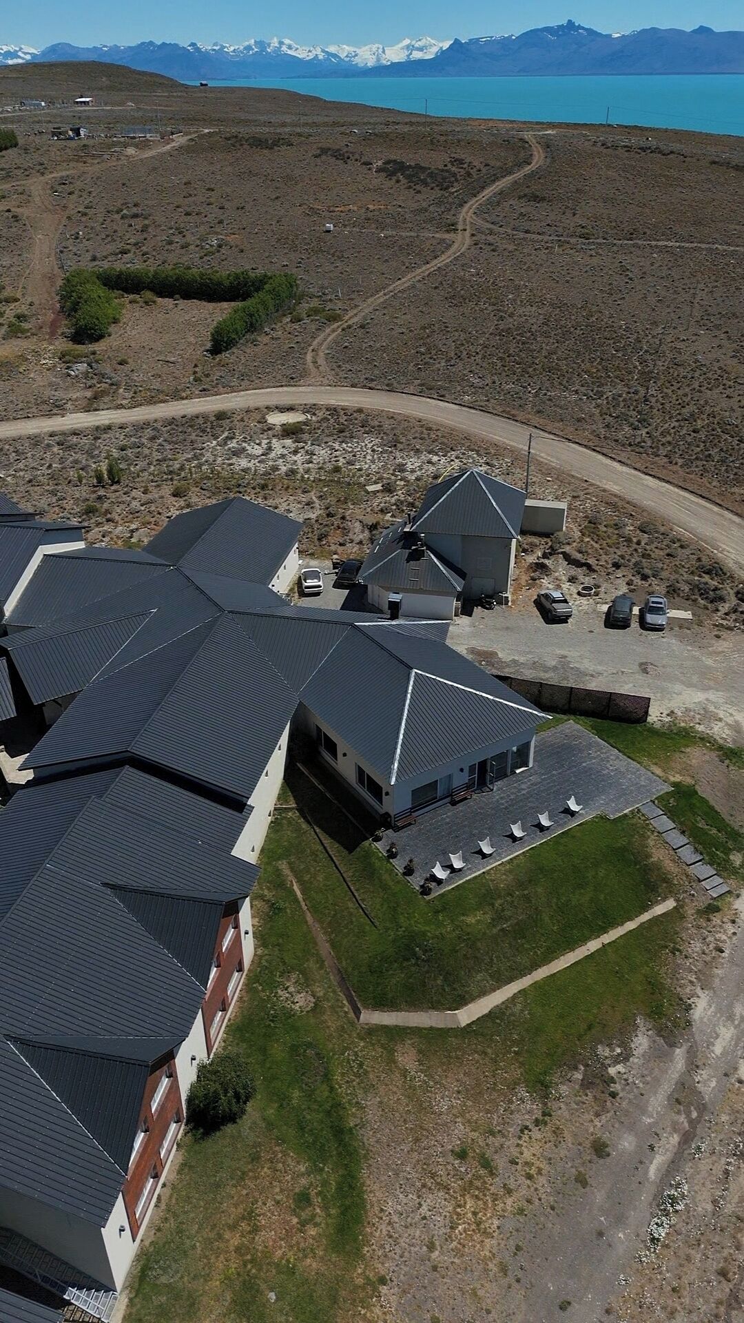 An aerial view of a residential area with multiple houses, a parking area, and a road leading to a body of water with mountains in the background.