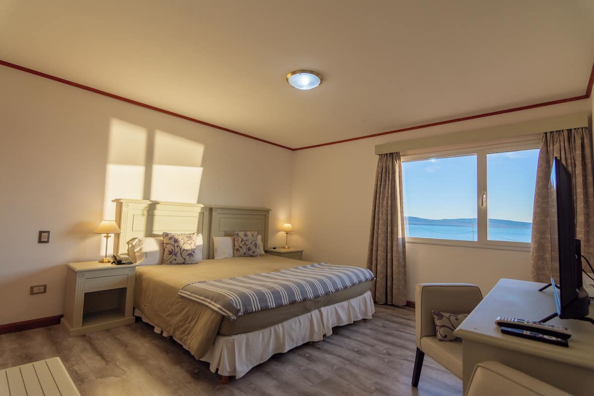 Quarto de hotel com cama, mesas de cabeceira, escrivaninha com cadeira, televisão e janela com cortinas.