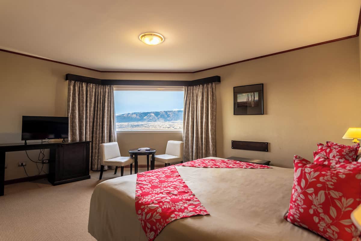 Quarto de hotel com cama, escrivaninha com televisão, cadeira, mesa pequena e janela com cortinas.