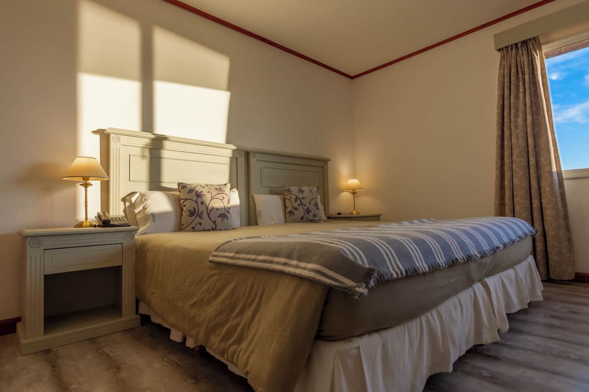 Quarto com cama, duas mesas de cabeceira, luminárias e janela com cortinas.