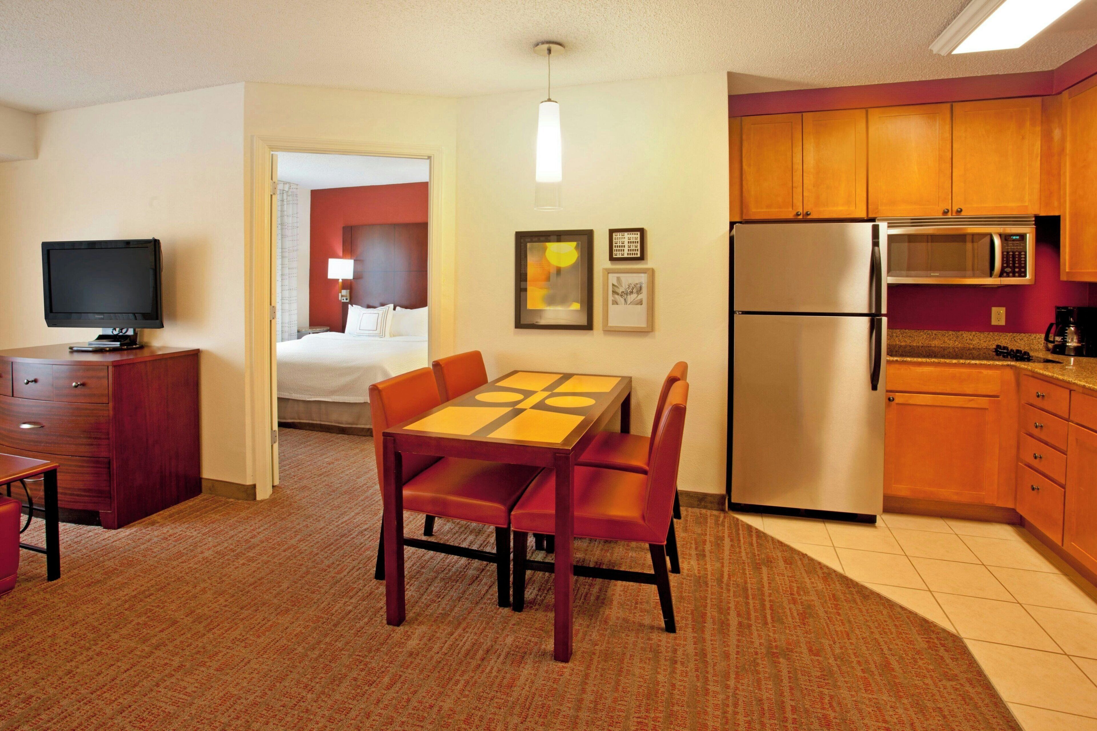 A hotel room with a dining table, chairs, a TV, a bed, a kitchen area with a refrigerator and microwave, and a carpeted floor.