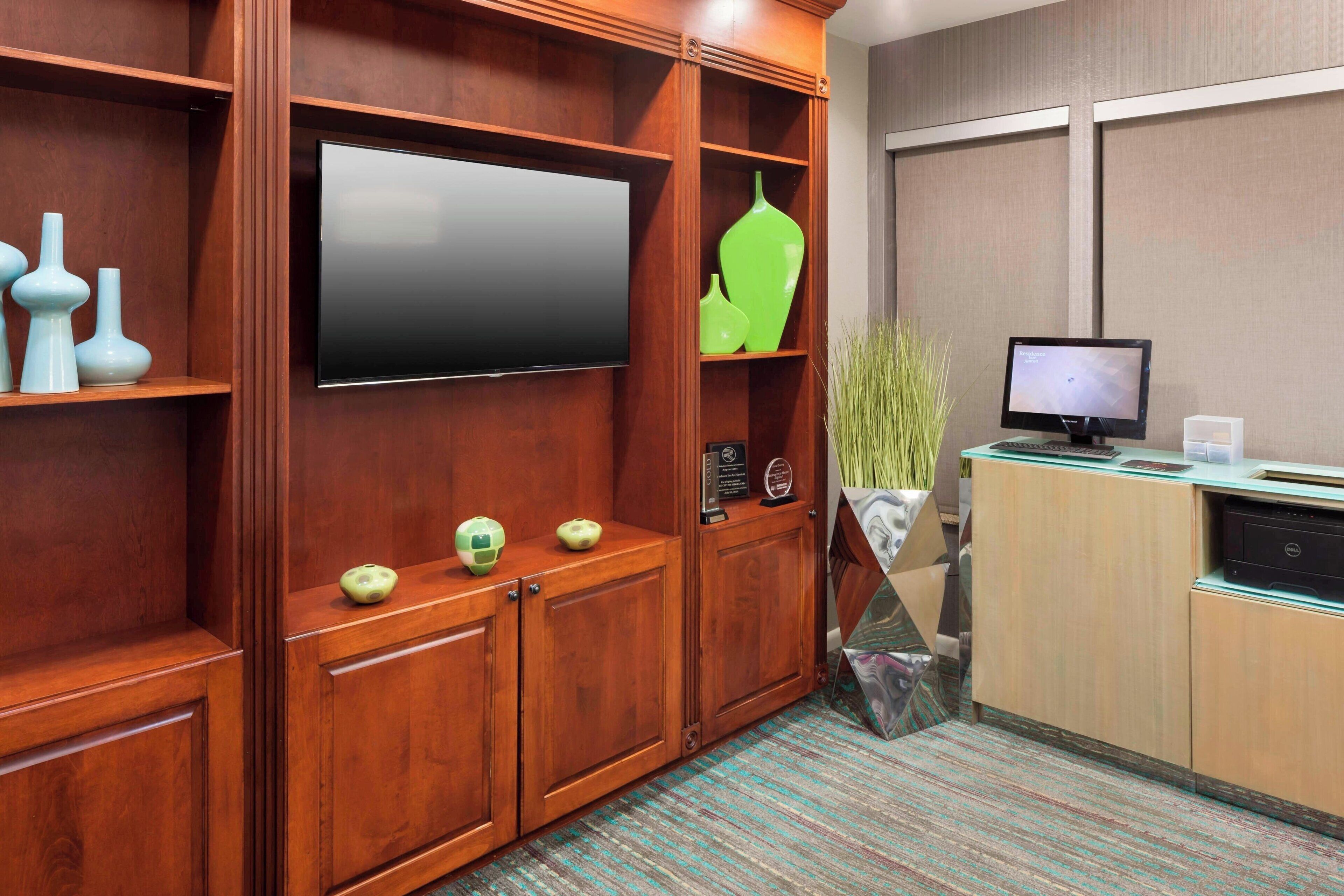 Quarto de hotel com um armário de madeira, uma televisão de tela plana, uma pequena mesa de escritório com um computador e uma planta.