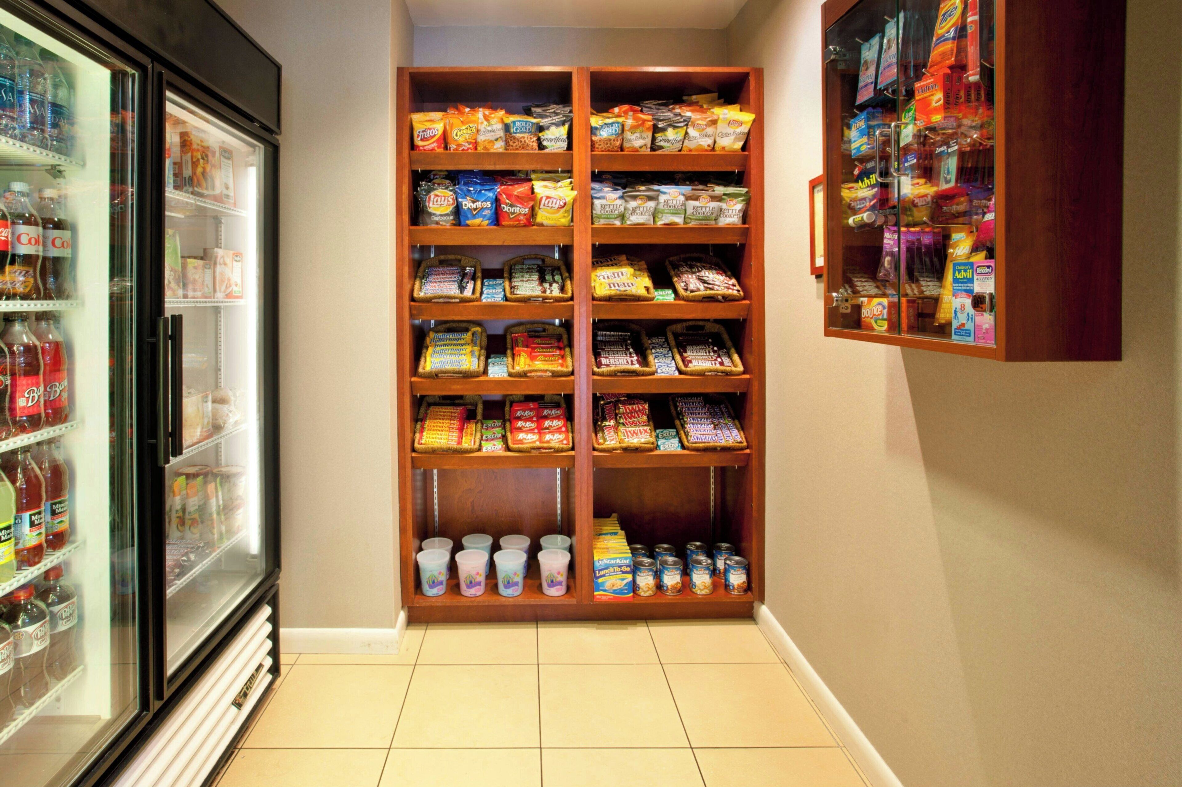 A snack and beverage display in a hotel corridor, including a refrigerated section with drinks and a wooden shelf with snacks and canned goods.