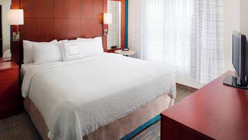 A hotel room with a large bed, a wooden headboard, a bedside table with a lamp, a small round table, and a TV on a wooden stand.
