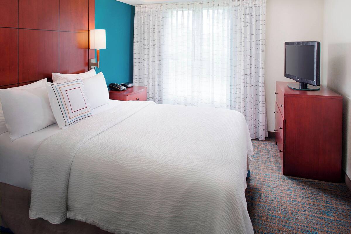 Habitación de hotel con una cama grande, una mesita de noche con lámpara, un televisor sobre un mueble de madera y una ventana con cortinas translúcidas.
