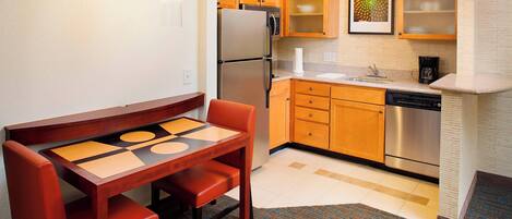A compact kitchen with wooden cabinets, a stainless steel refrigerator, a dishwasher, and a dining table with two chairs.