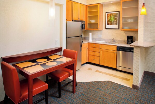 A compact kitchen with wooden cabinets, a stainless steel refrigerator, a dishwasher, and a dining table with two chairs.