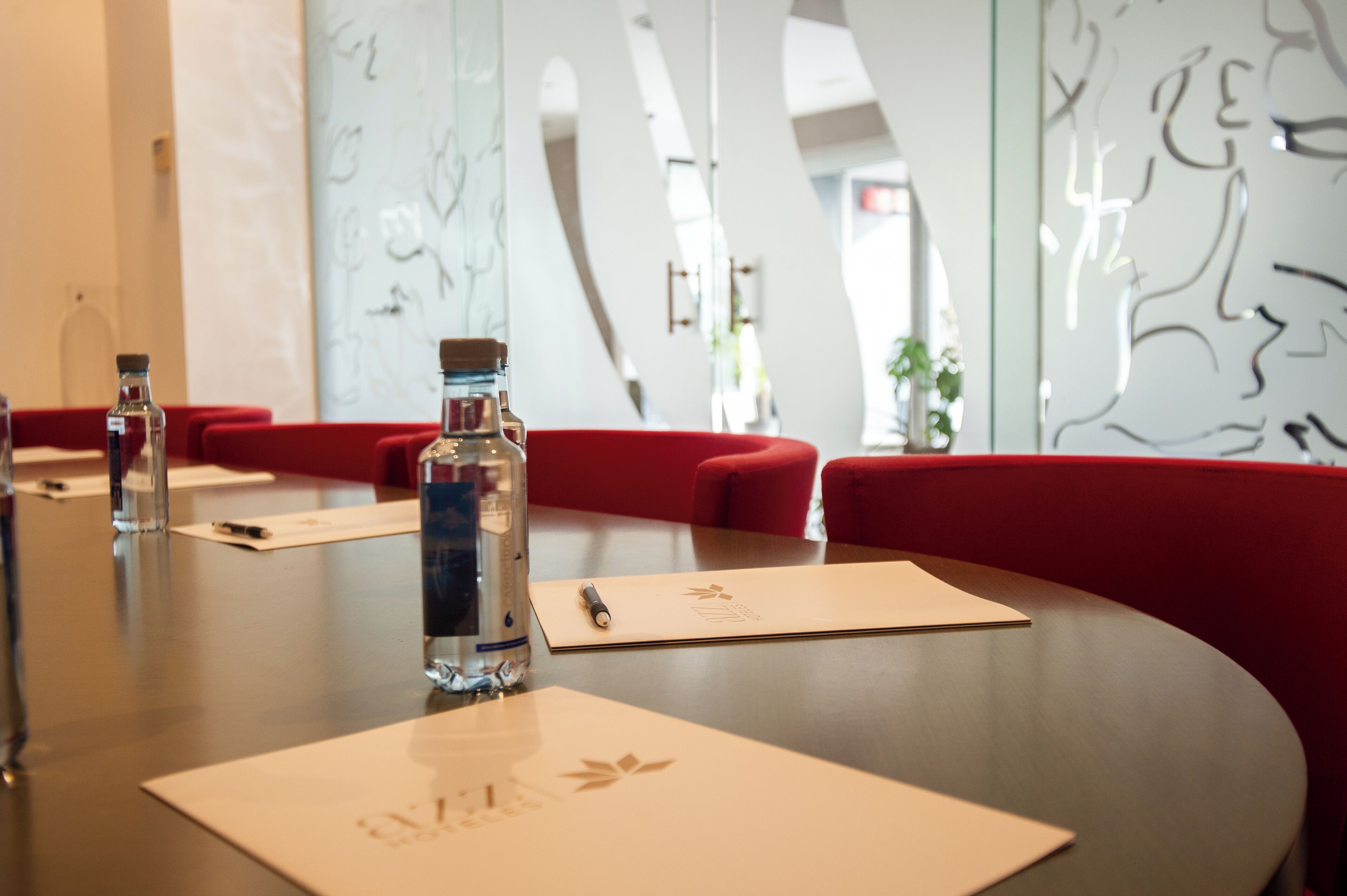 A conference table with water bottles, name cards, and red upholstered chairs in a modern interior with abstract wall art.