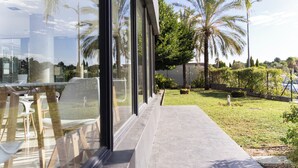 Um edifício moderno com grandes janelas que dão para um jardim bem cuidado, com palmeiras e um céu limpo.