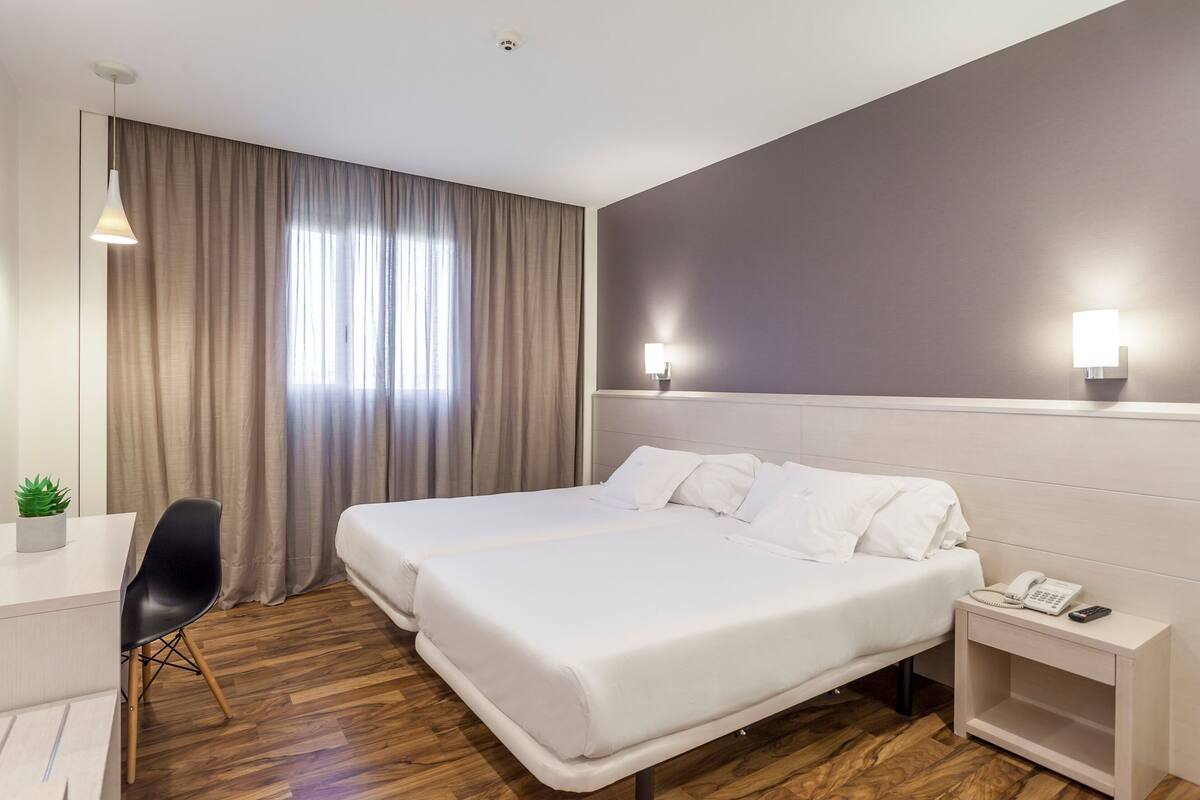A hotel room with two beds, a desk, a chair, a small plant, and a nightstand with a phone.