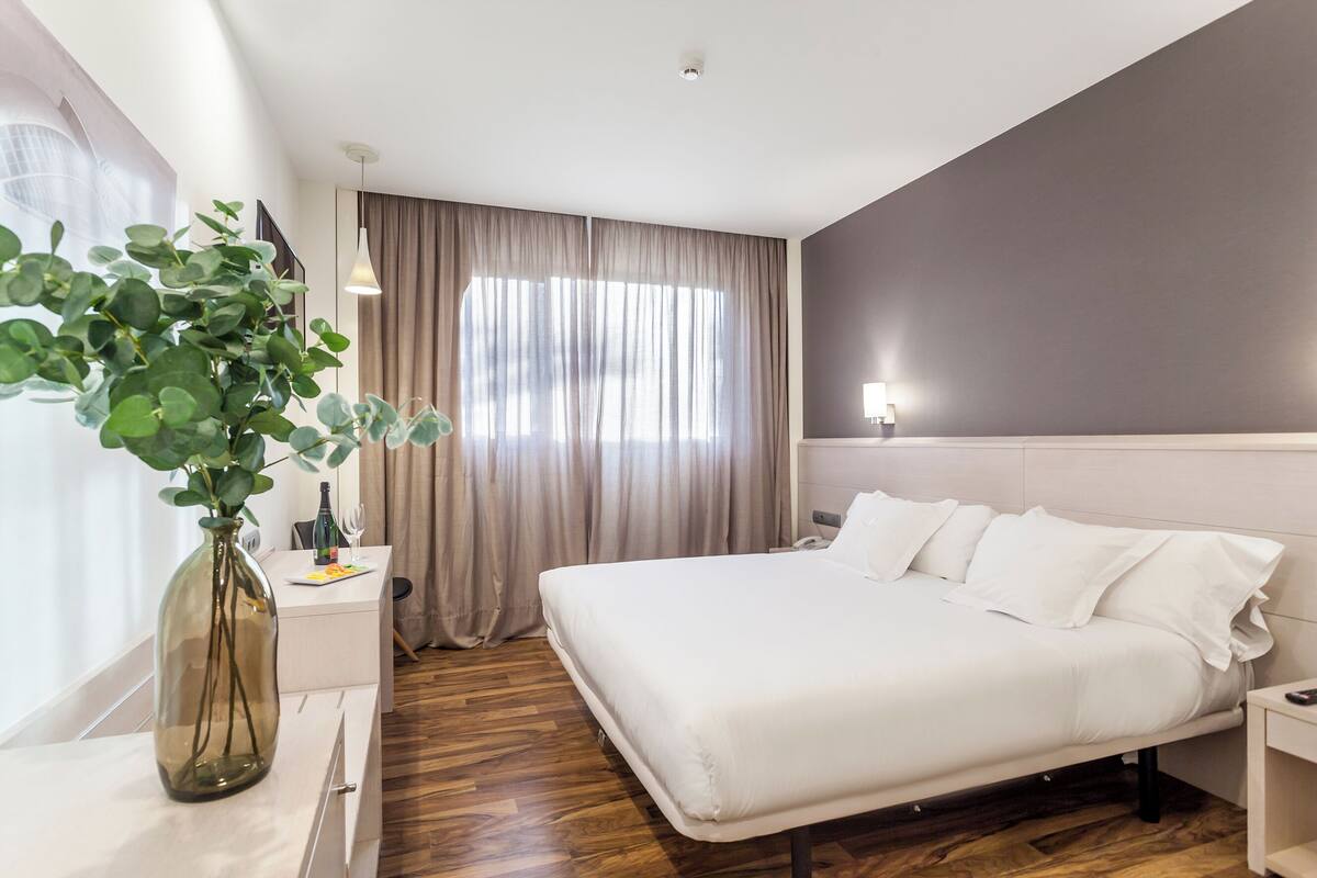 A modern hotel room with a large bed, a bedside table, a nightstand, a potted plant, and a window with curtains.