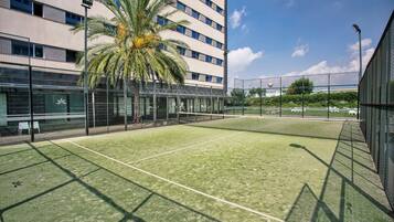 Cancha de tenis al aire libre con una palmera, rodeada por un edificio moderno con paredes de vidrio y una valla.