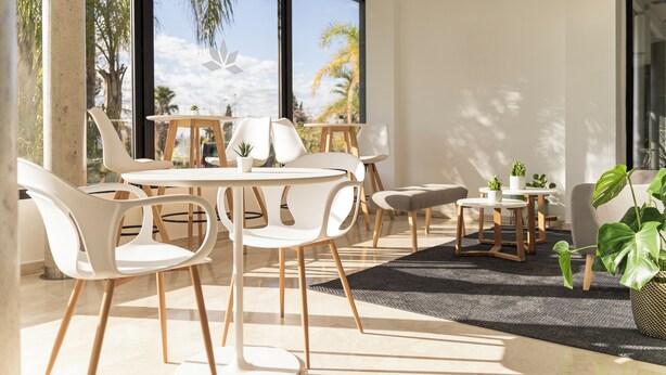 Um interior moderno com cadeiras e mesas brancas, grandes janelas com vista para palmeiras e um vaso de planta.