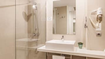 Un baño moderno con ducha de vidrio, lavamanos blanco, espejo y secador de pelo en la pared.