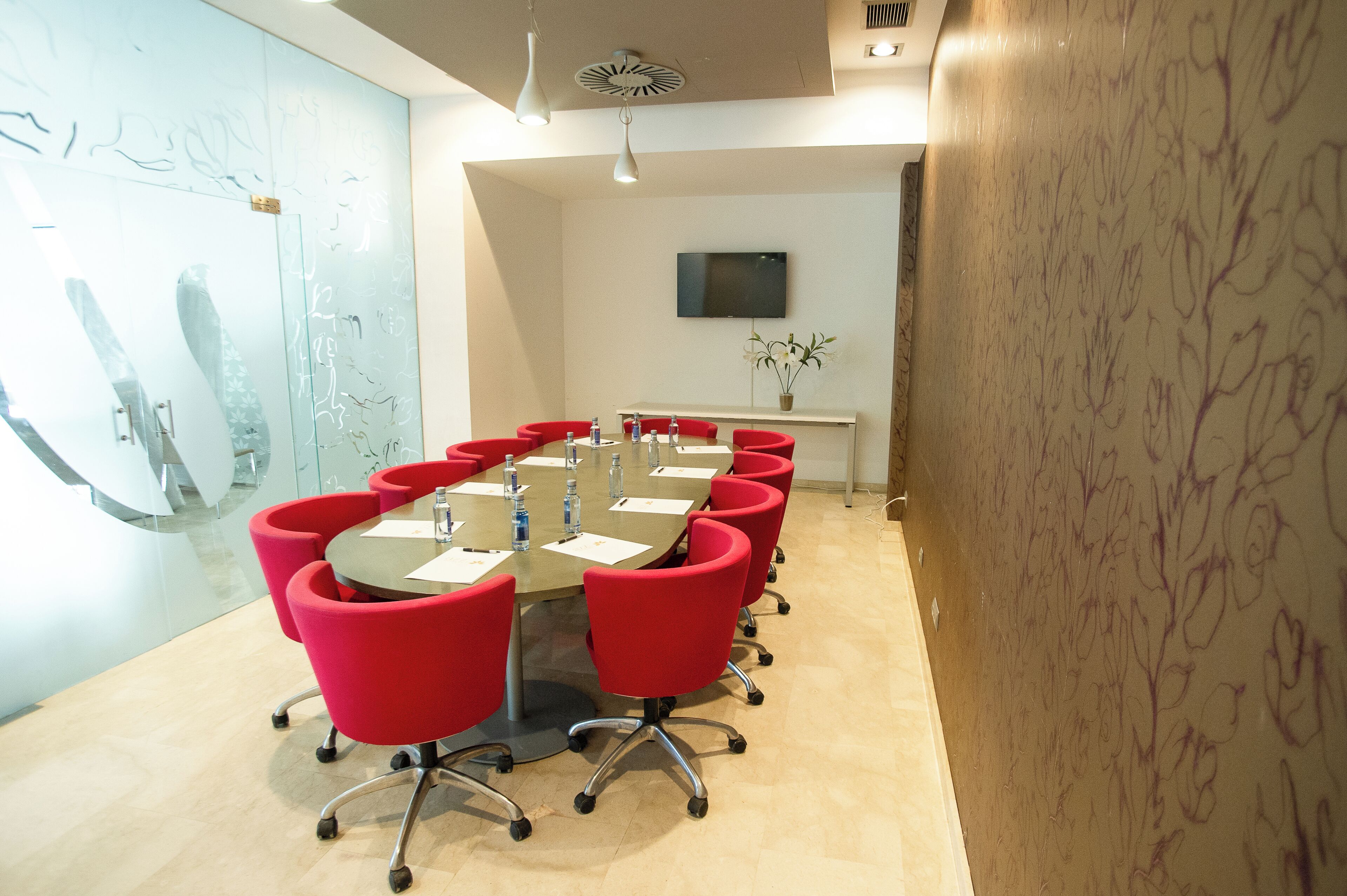 A conference room with a long table, red chairs, a flat-screen TV, and a potted plant.