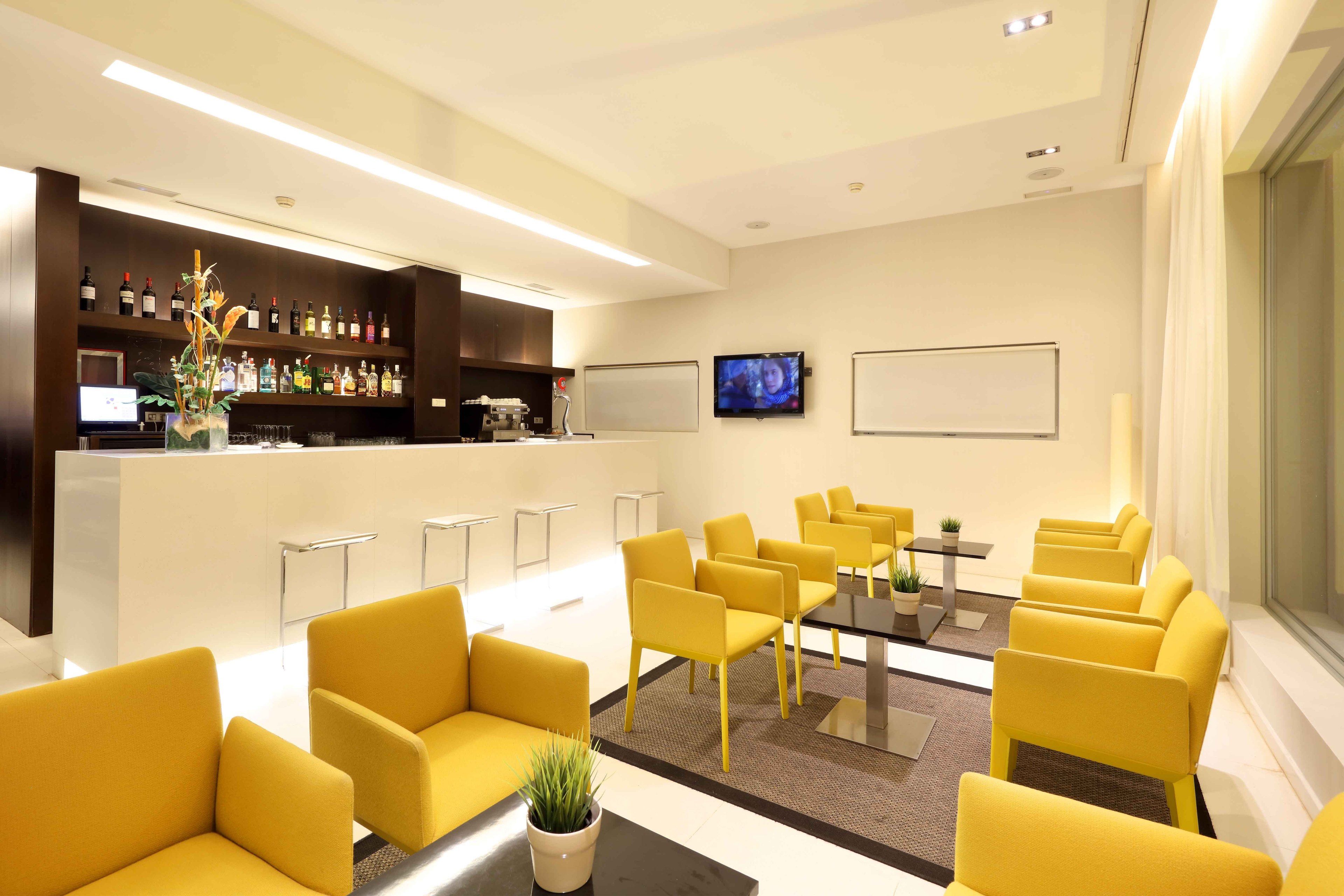 A modern lobby with yellow seating, a bar area with various bottles, a TV, and a whiteboard.