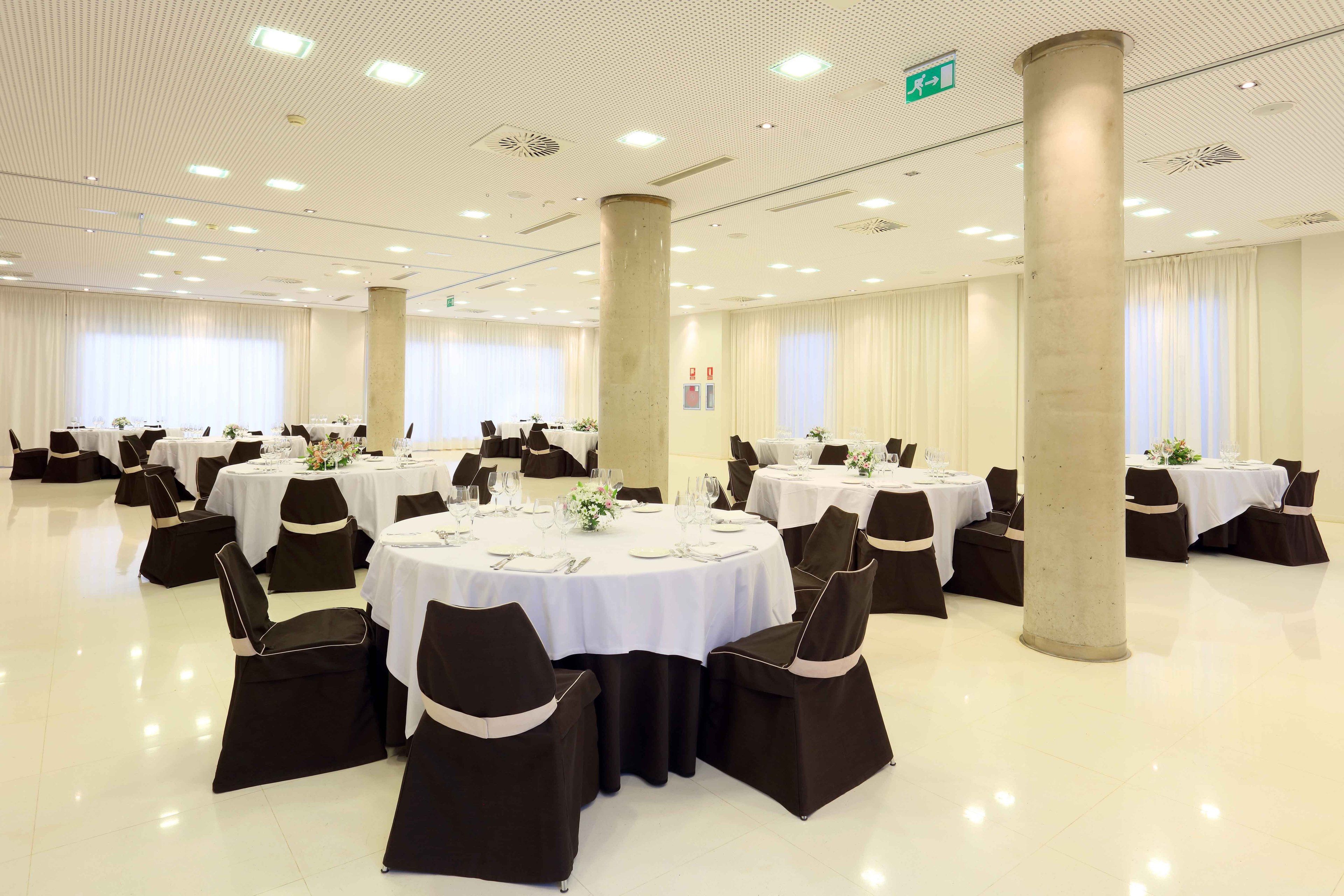 Un amplio salón de banquetes bien iluminado con mesas redondas dispuestas para comer, sillas negras con cintas blancas y un diseño de techo moderno y limpio.