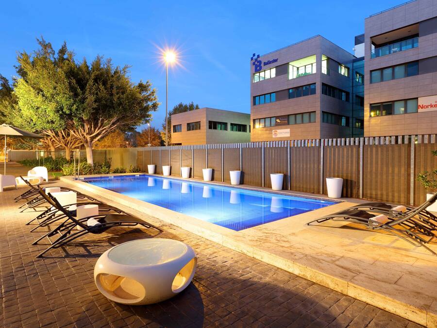 Una piscina con sillones de descanso y una zona de estar hundida, rodeada de una valla y edificios modernos al fondo.