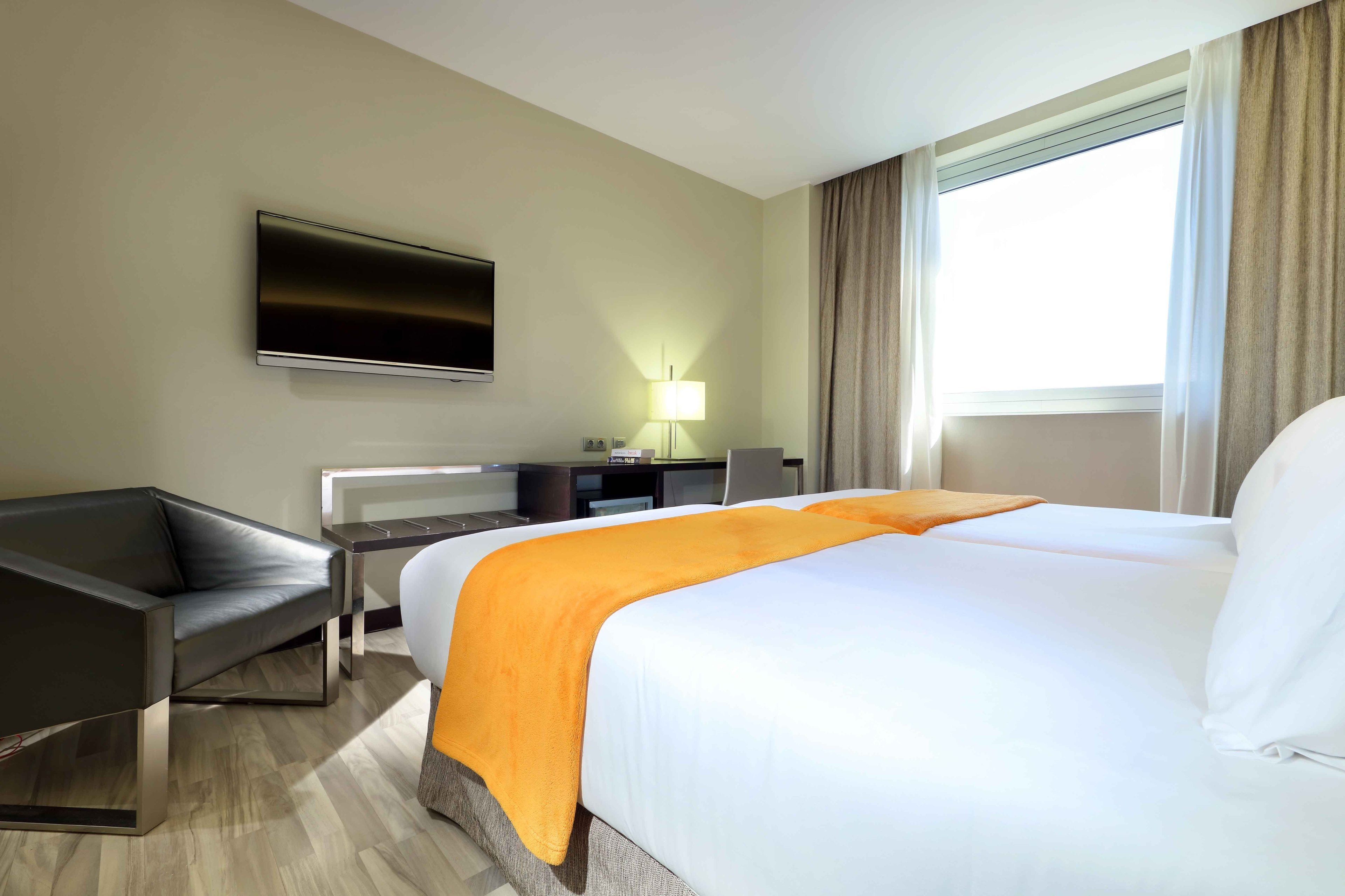 A hotel room with a large bed, a desk with a chair, a flat-screen TV mounted on the wall, and a window with curtains.
