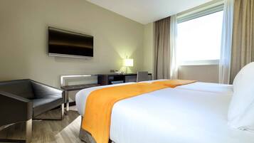 A hotel room with a large bed, a desk with a chair, a flat-screen TV mounted on the wall, and a window with curtains.