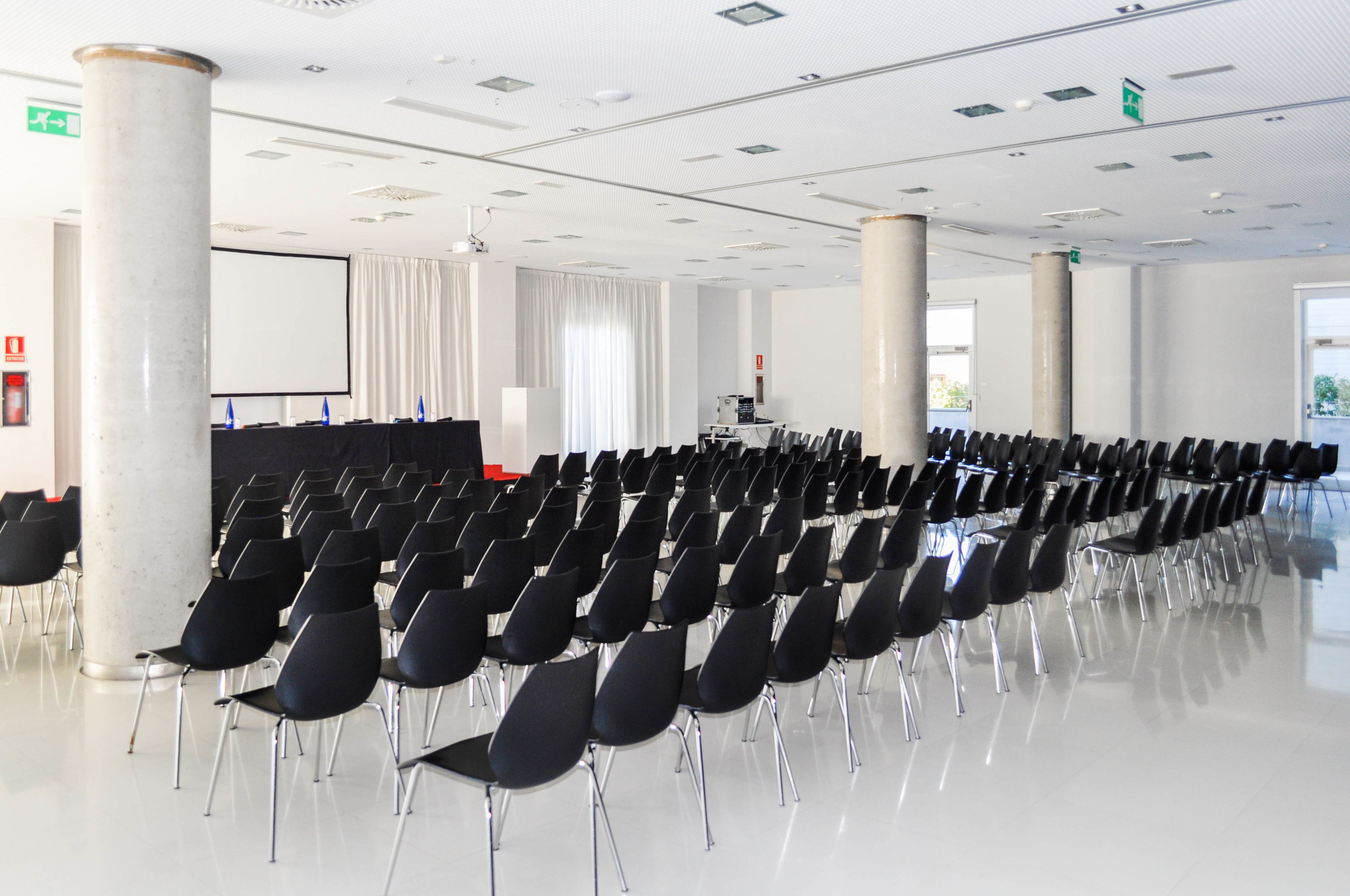 Un salón de conferencias con hileras de sillas negras, una pantalla grande y un atril.