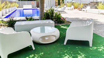 A poolside area with white lounge chairs and a glass-top table on artificial grass, surrounded by potted plants and a blue pool.