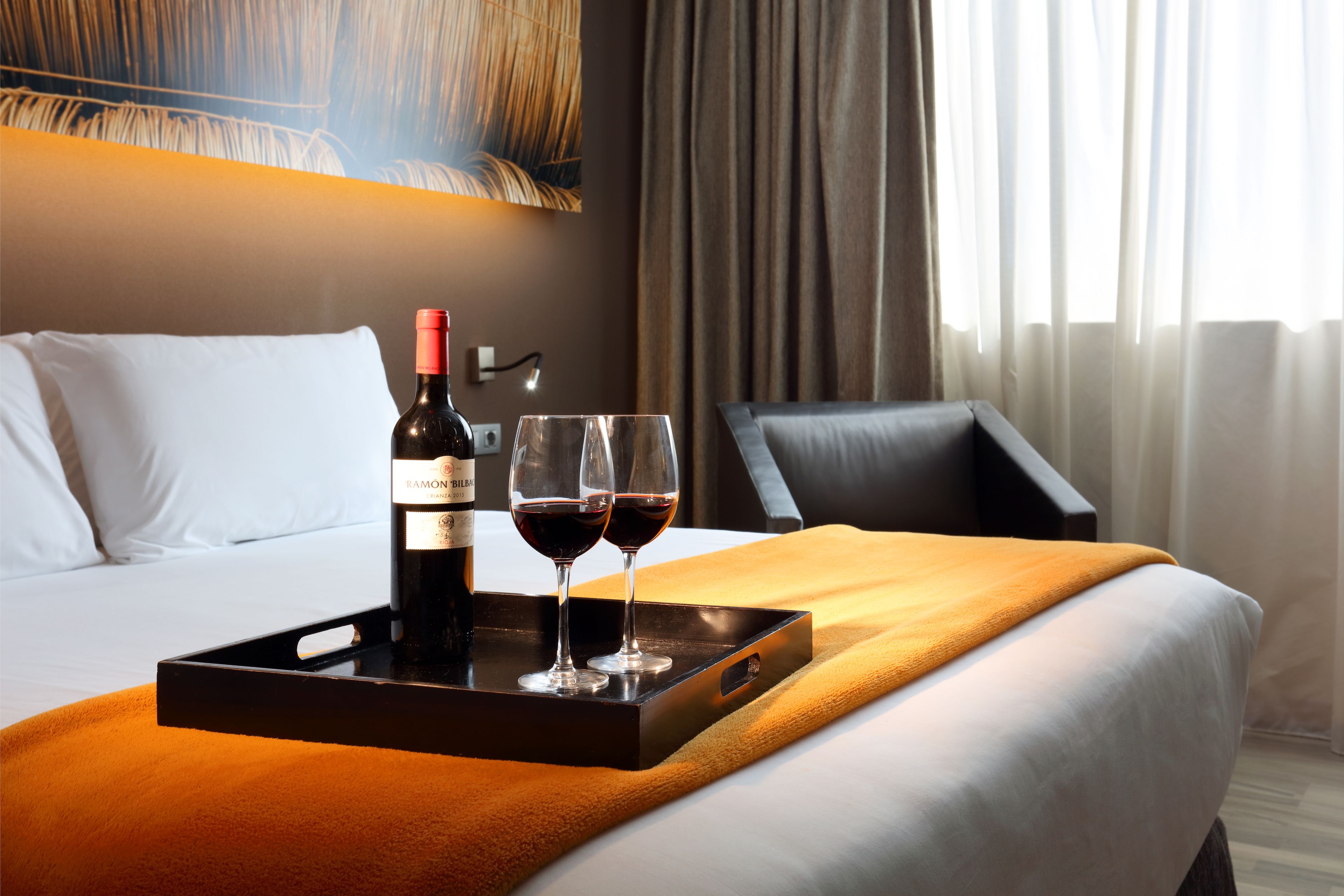 Une chambre d’hôtel avec un lit, un plateau contenant une bouteille de vin et deux verres, et une chaise.