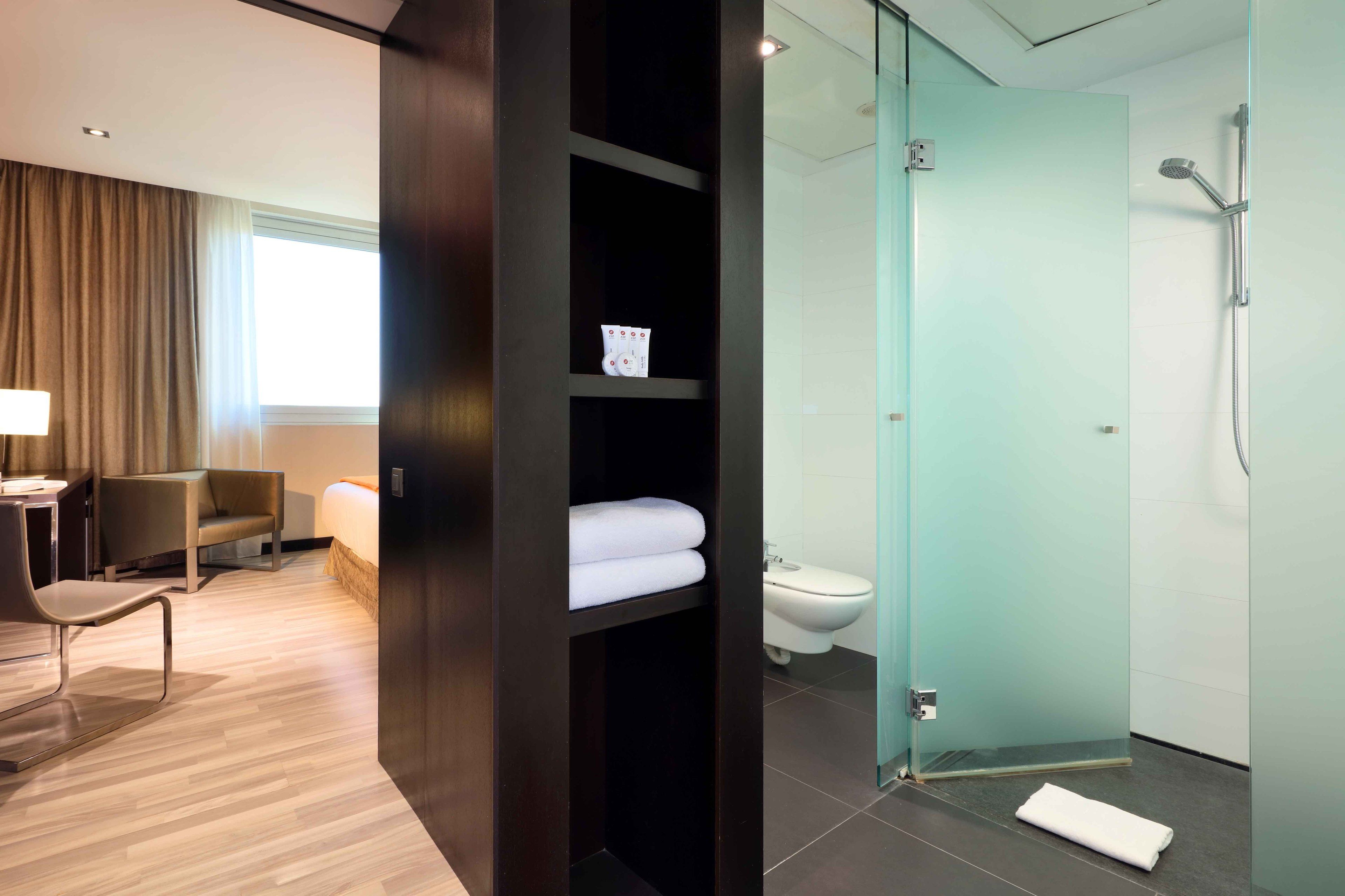 Ein modernes Hotelzimmer mit Holzboden, Schreibtisch mit Stuhl, Bett, einer Duschkabine aus Glas sowie einer Toilette.