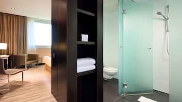 Ein modernes Hotelzimmer mit Holzboden, Schreibtisch mit Stuhl, Bett, einer Duschkabine aus Glas sowie einer Toilette.