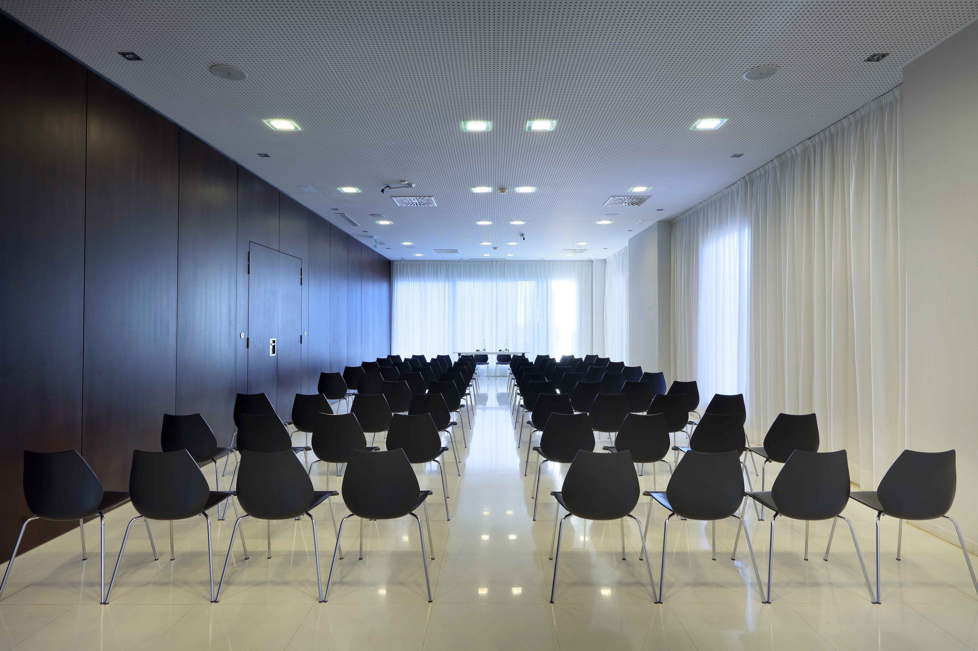 A conference room with rows of chairs, a white ceiling with recessed lighting, and large windows with curtains.