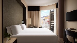 A hotel room with a large bed, a desk, a chair, a TV, and a window with a view of buildings.