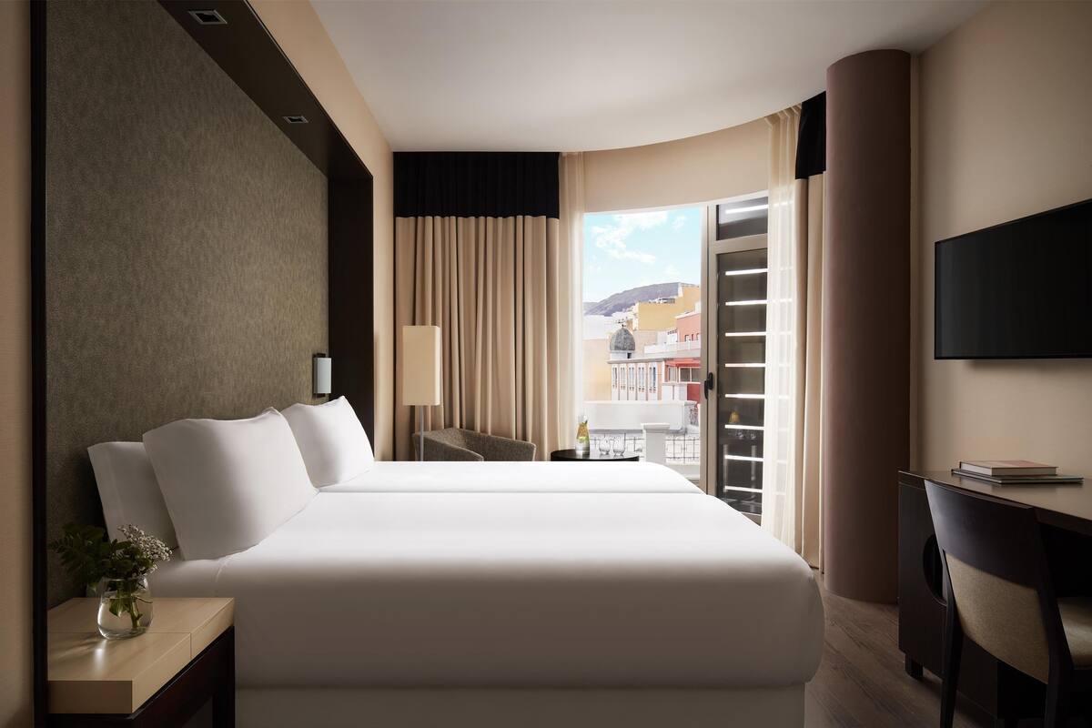 Une chambre d’hôtel avec un grand lit, un bureau, une chaise, une télévision et une fenêtre donnant sur des immeubles.