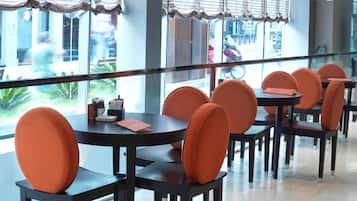 Un restaurante con mesas redondas y sillas anaranjadas, ventanas amplias con persianas y vista a la calle.