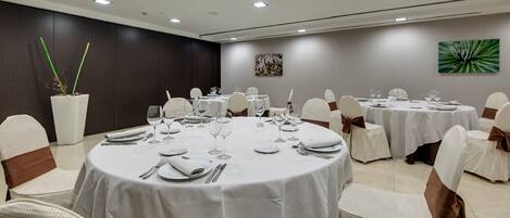 Un salón de conferencias con mesas redondas, manteles blancos y sillas blancas adornadas con fajas marrones.