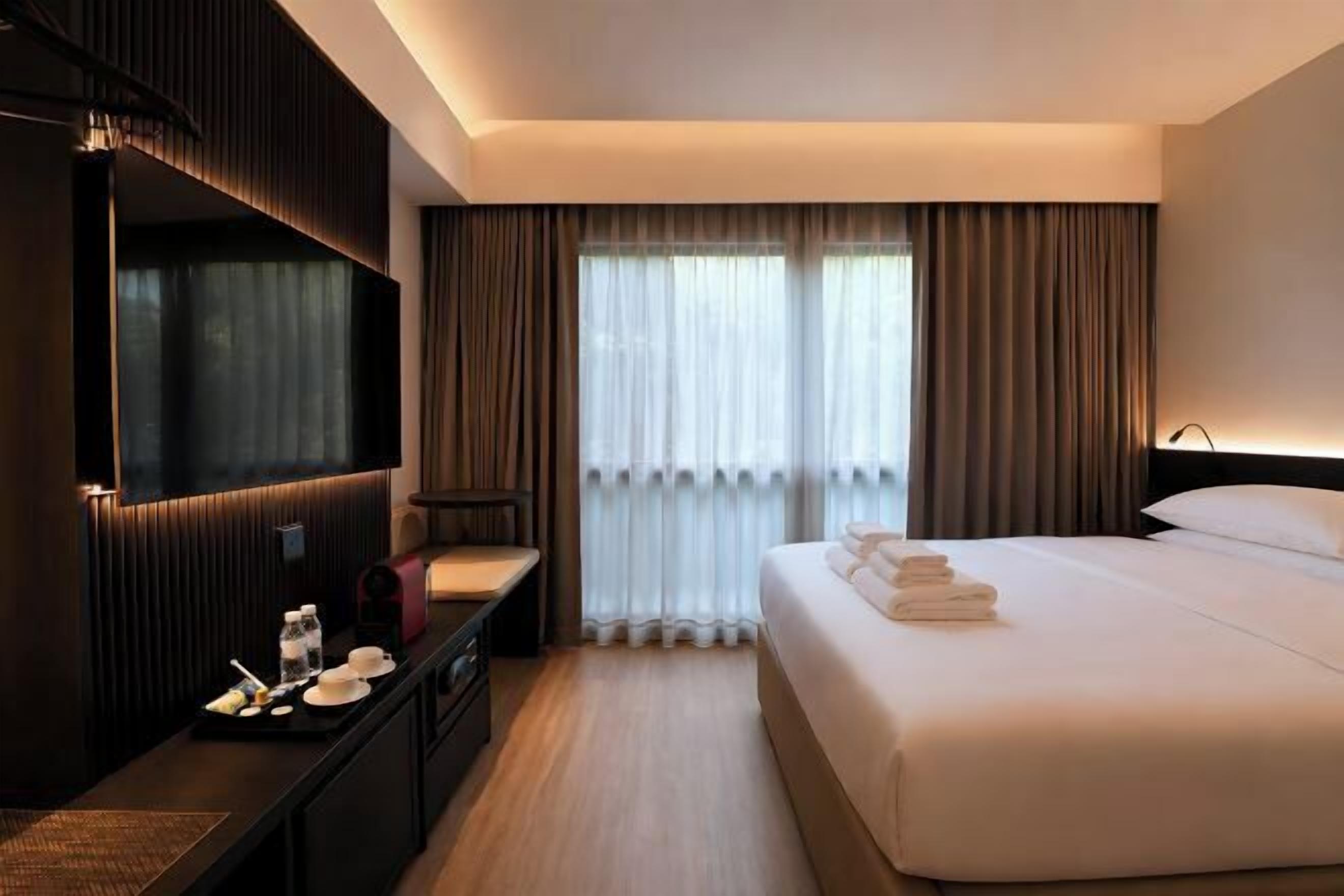 A hotel room with a bed, a desk with a chair, a TV, and a large window with curtains.