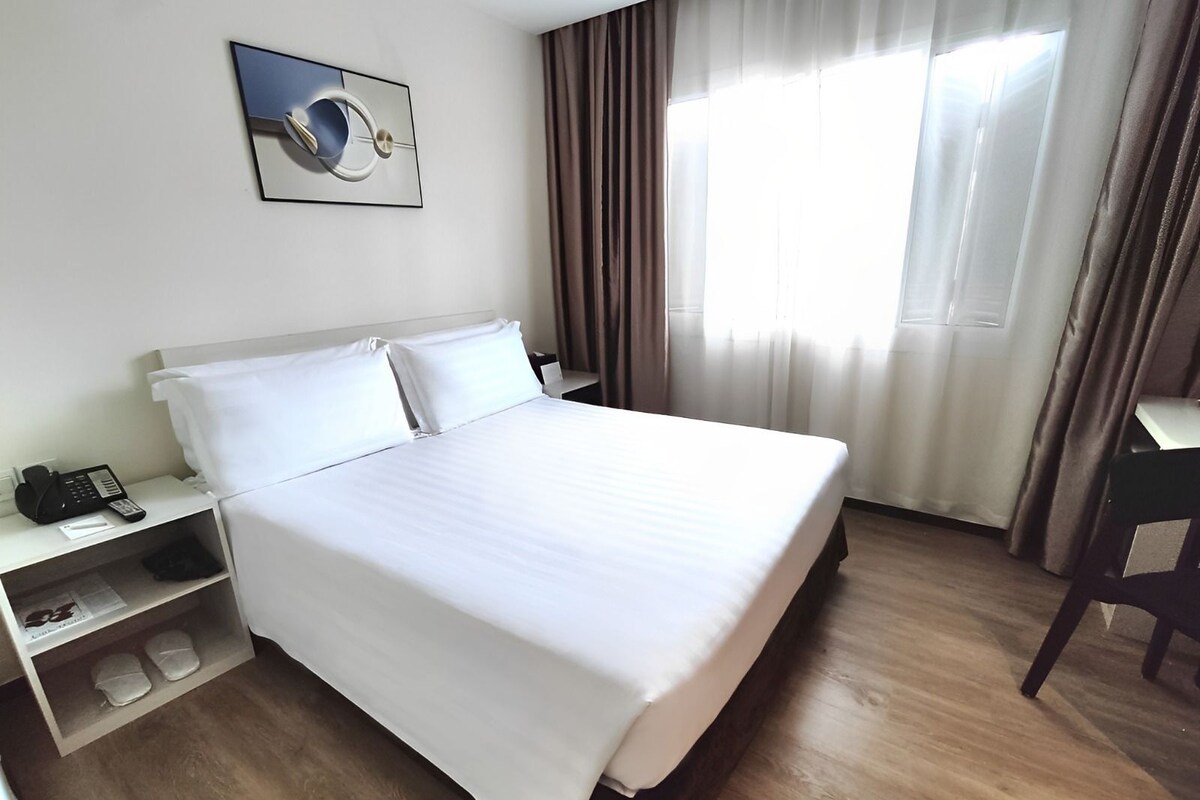A hotel room with a bed, bedside table with a phone, a desk with a chair, and a window with curtains.