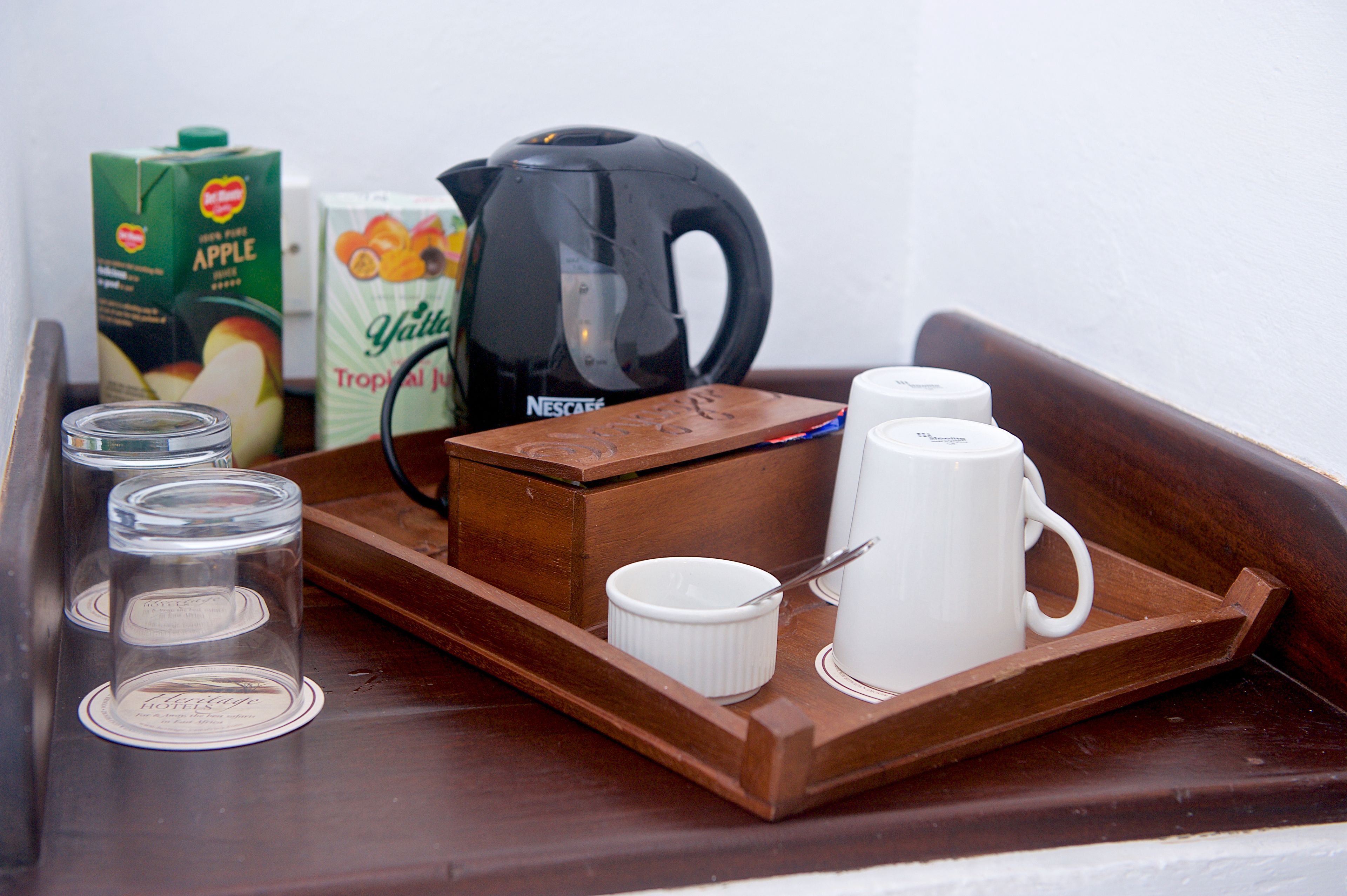 A wooden tray with a Nescafé coffee maker, a carton of apple juice, a carton of tropical juice, and three clear glass cups.