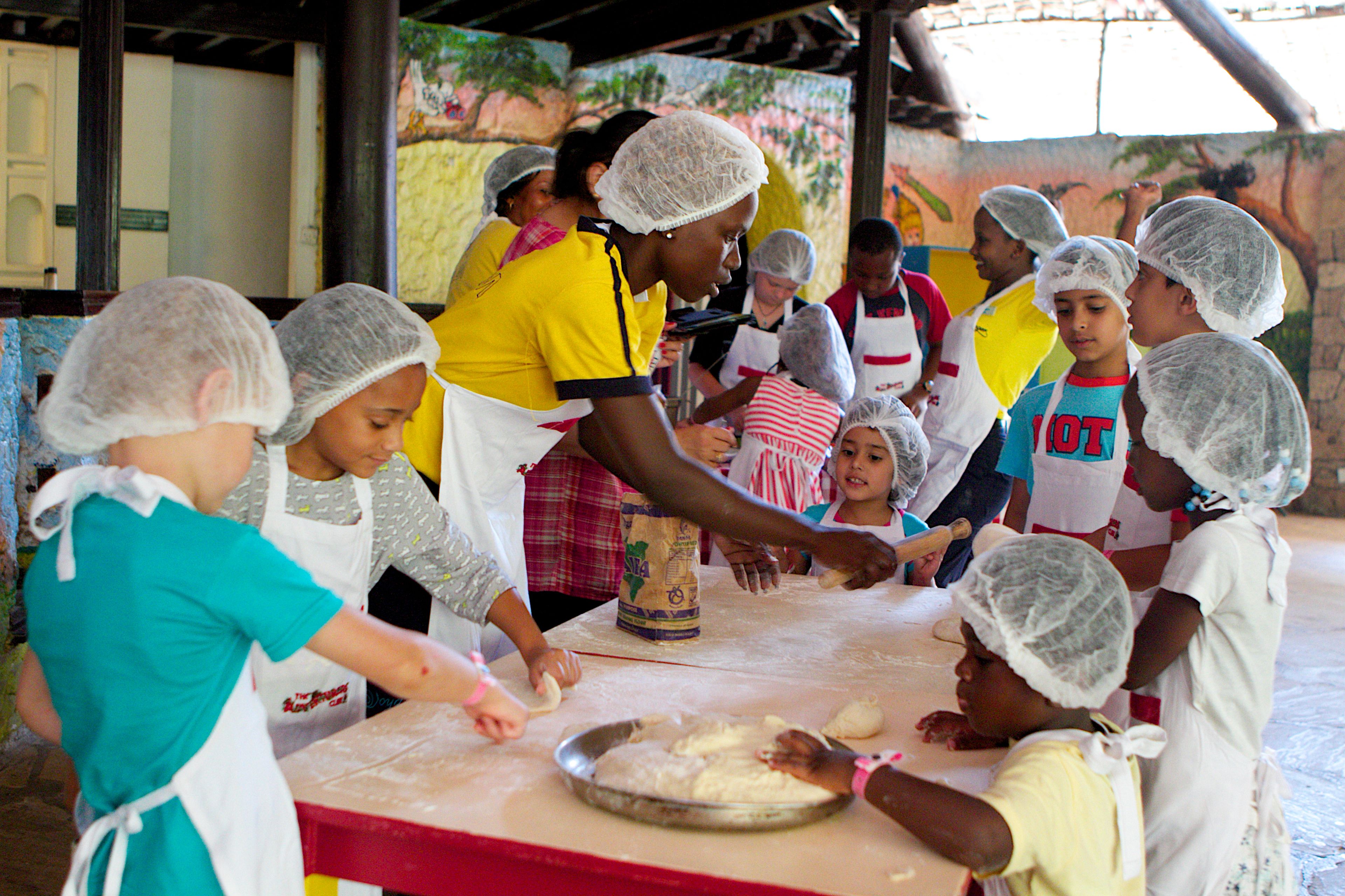 A group of children and adults engaged in a baking activity, wearing hairnets and aprons.