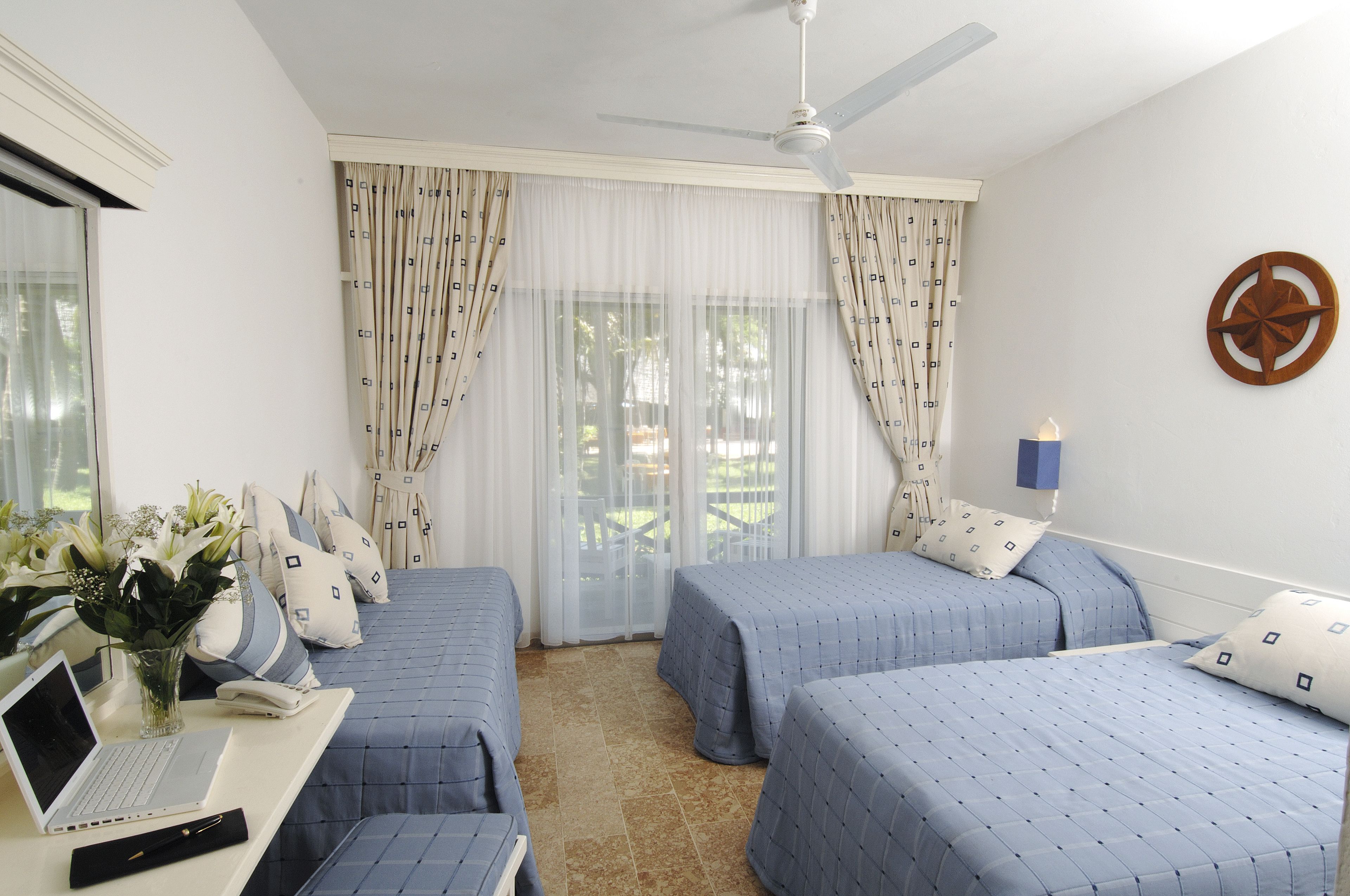 A hotel room with two beds, a desk with a laptop, a vase with flowers, and a ceiling fan.