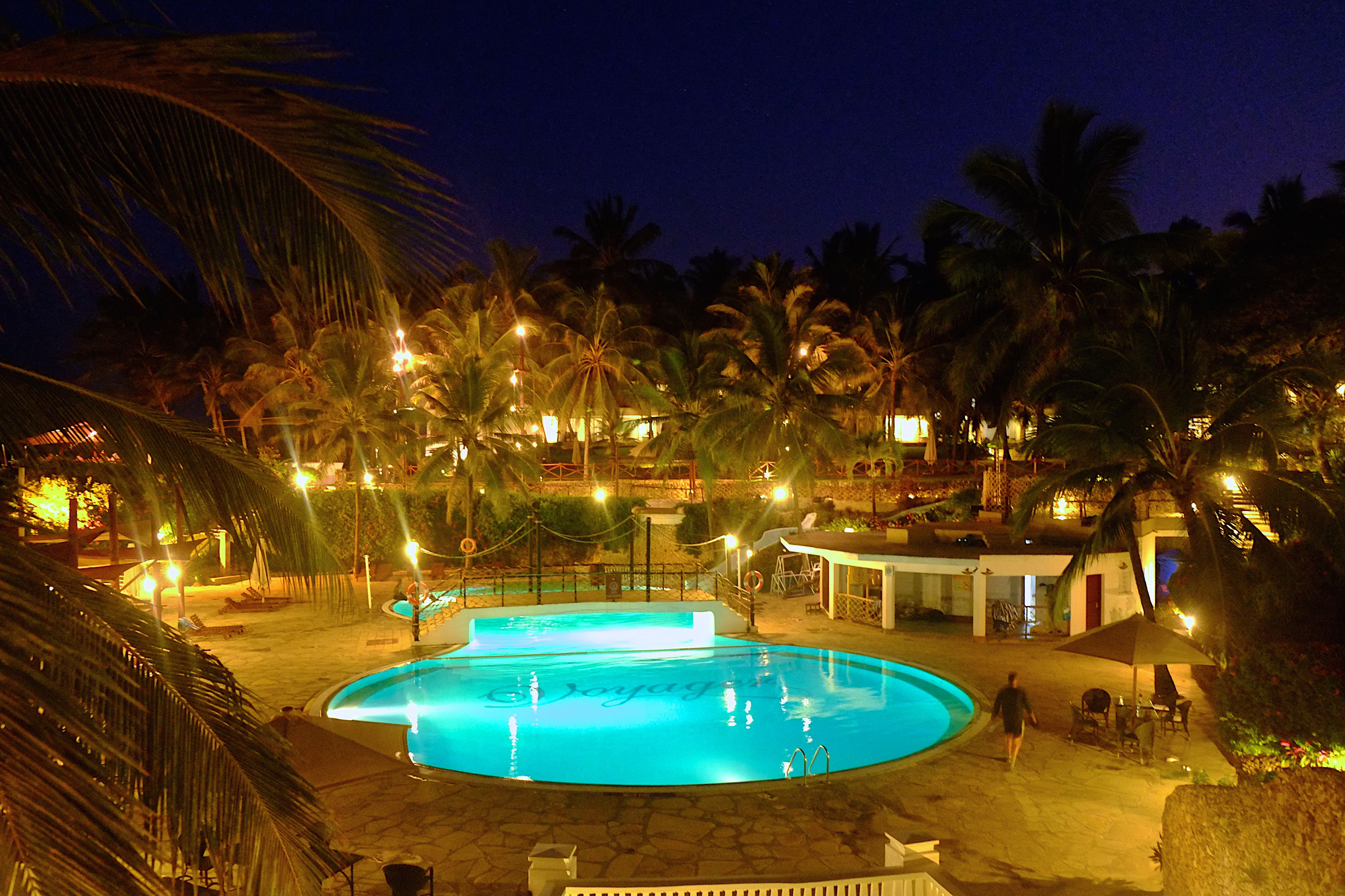 Vista noturna de uma área de piscina com palmeiras e um edifício ao fundo.