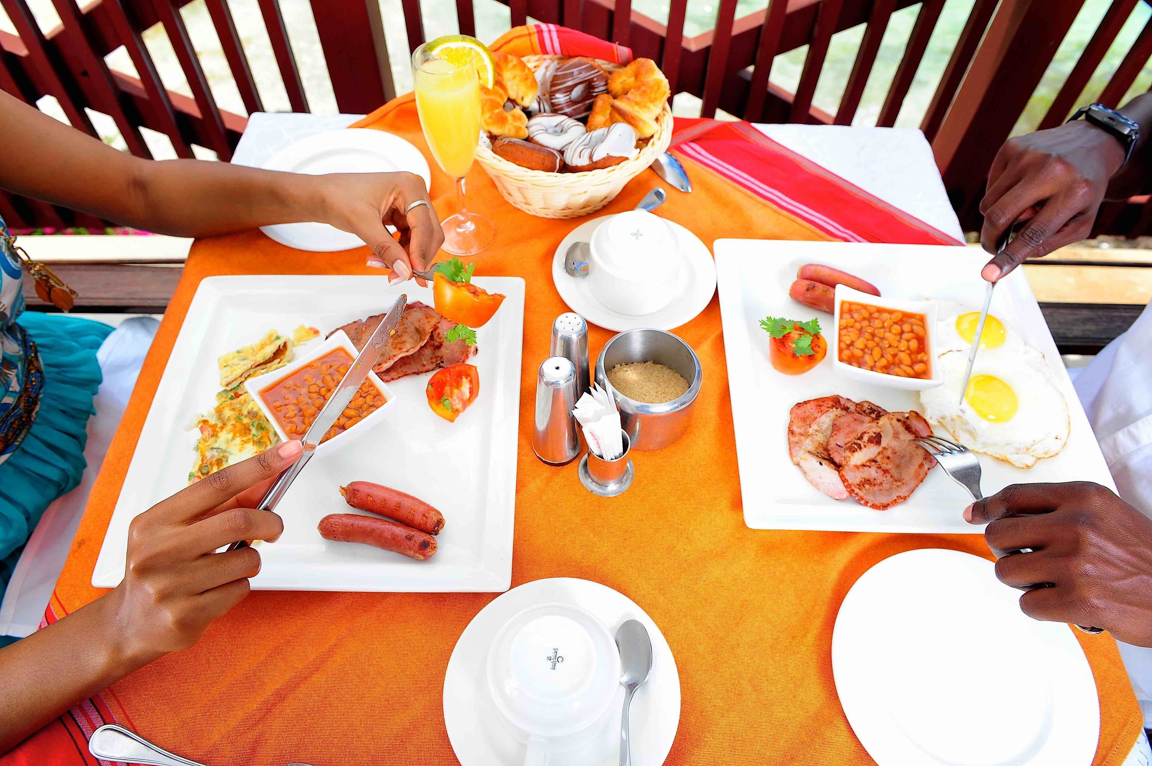 A group of people dining at an outdoor table with various breakfast items including eggs, sausages, and a fruit salad.