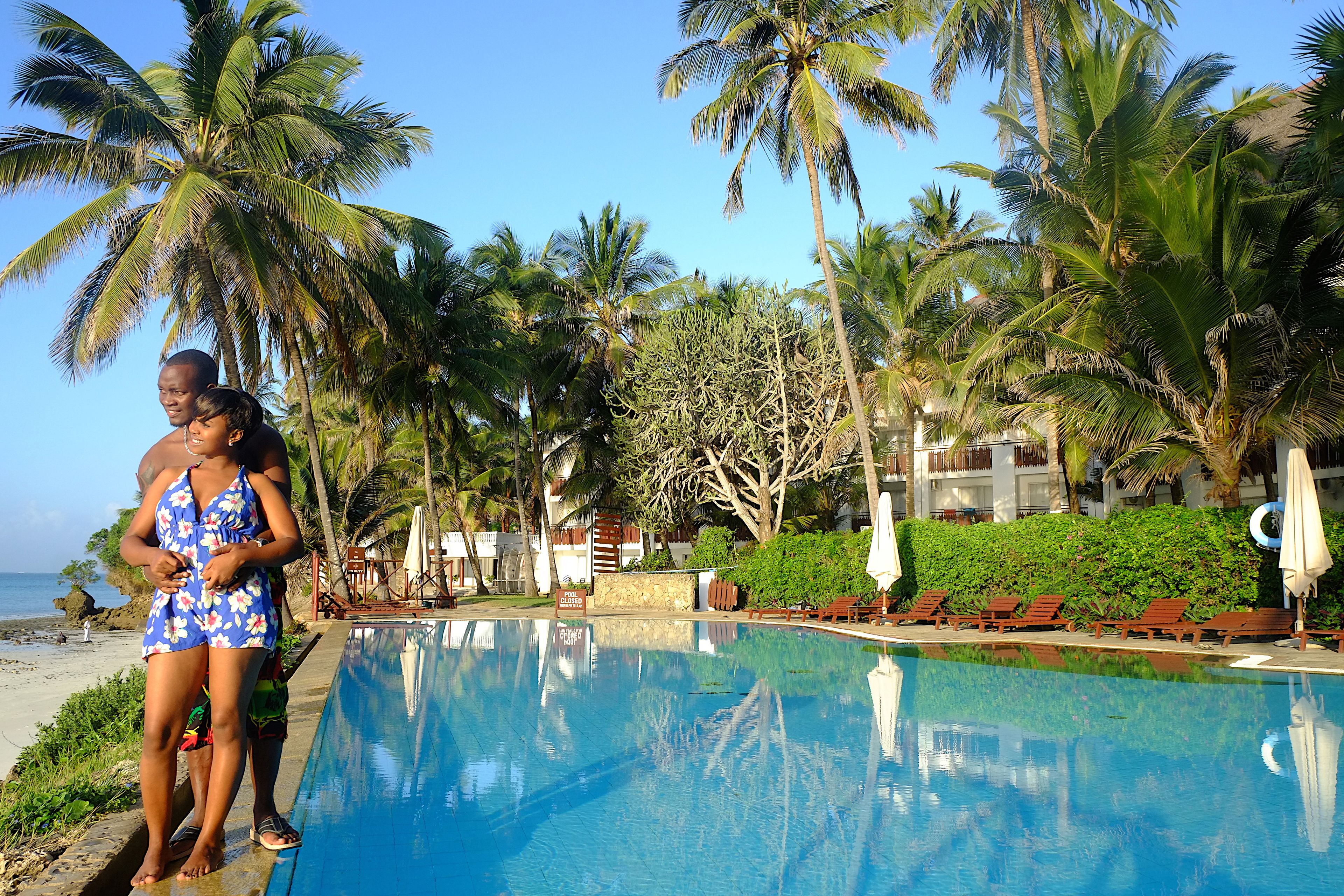 Zona junto a la piscina con palmeras, vista a la playa y sillas de descanso.