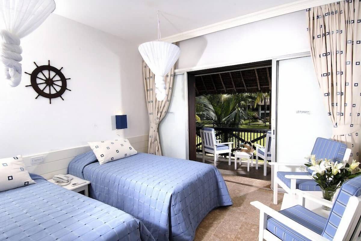 A hotel room with two beds, a small table, a chair, a balcony with a view of palm trees, and a ship's wheel decoration.