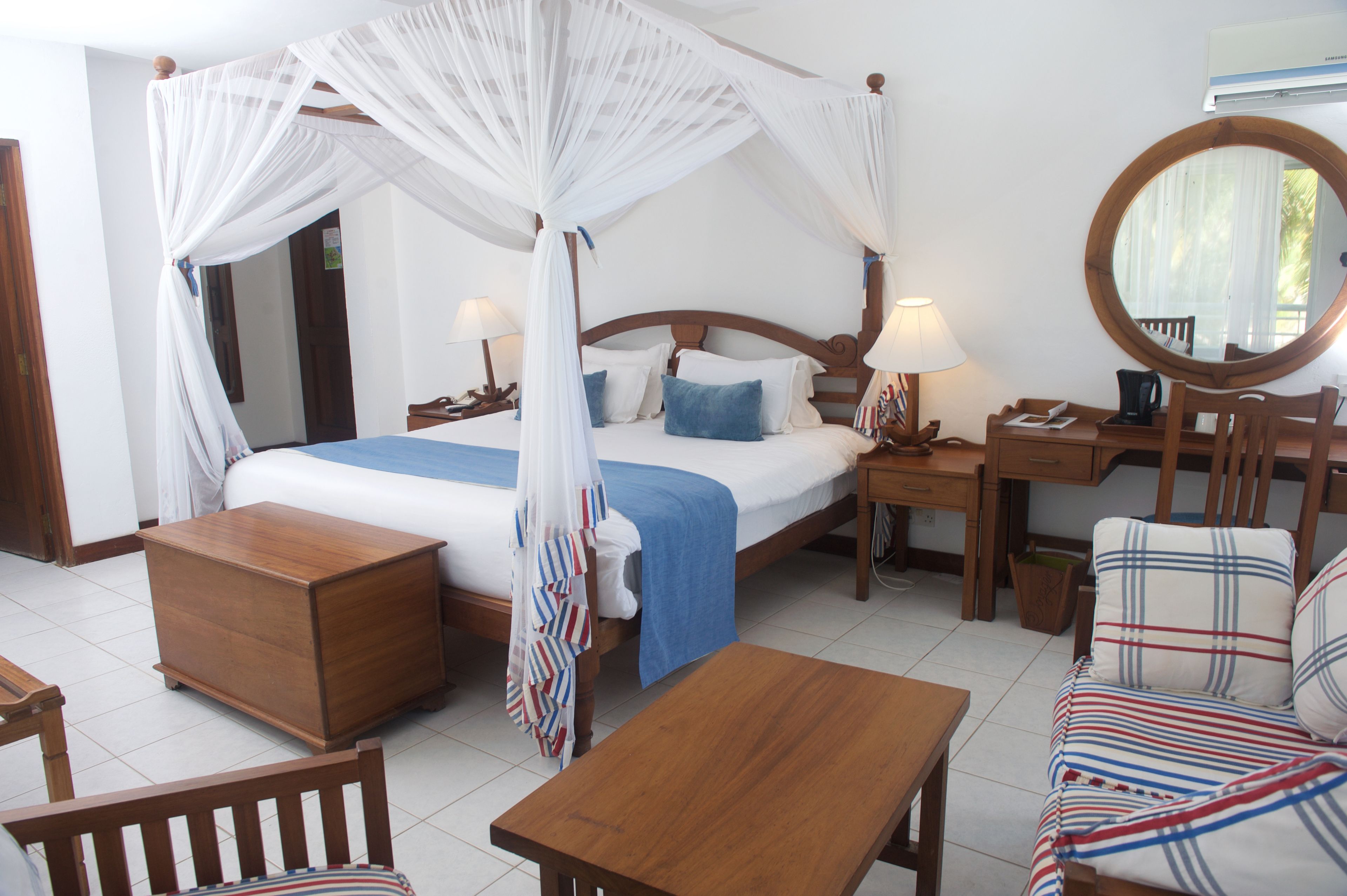 A bedroom with a four-poster bed, bedside tables with lamps, a wooden nightstand, a chair, a sofa, and a round mirror.