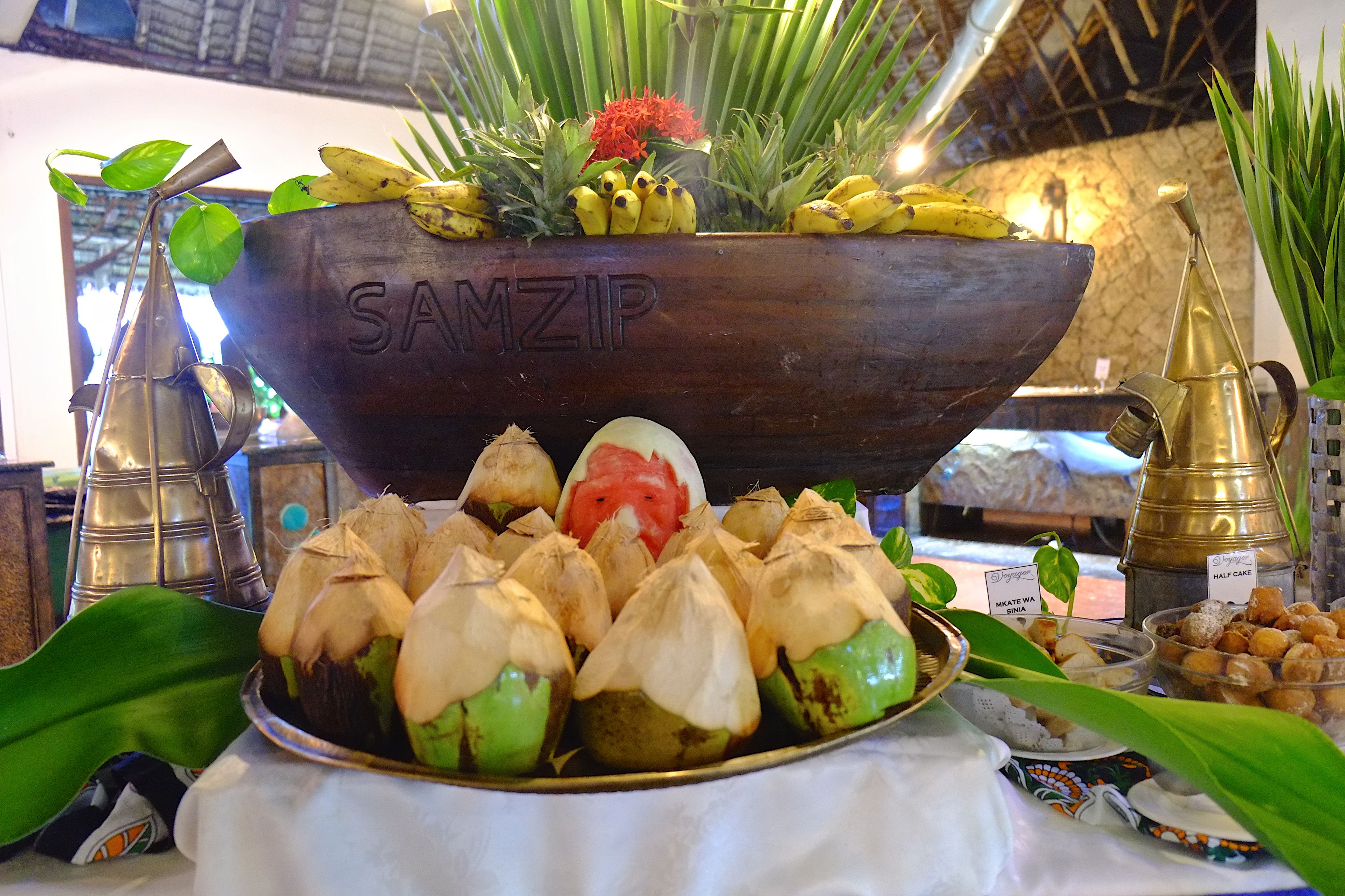 A display of traditional food items, including a large metal bowl labeled "SANZIP" and various dishes with food covered in leaves.